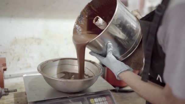 ワークショップでスケールのボウルに液体チョコレートを注ぐエプロンと手袋の匿名の女性の菓子作物の側面のビュー — ストック動画