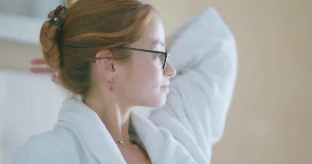 浅色卧室里穿着柔软的白色浴衣和眼镜 触摸着长长的波浪形姜黄头发的年轻女性千年 — 图库视频影像