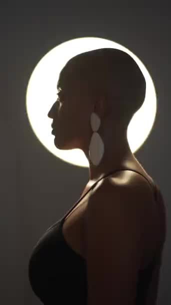 Side view of calm African American female with bald haircut and in trendy earrings standing in dark studio on background of round glowing lamp