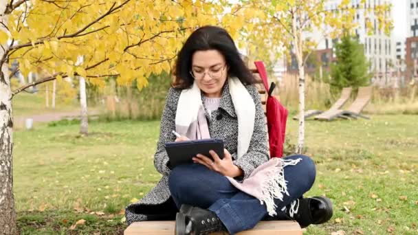 秋の公園に座っている間ベンチとブラウジングタブレットに座って毛布に包まれた幸せな若い女性 — ストック動画