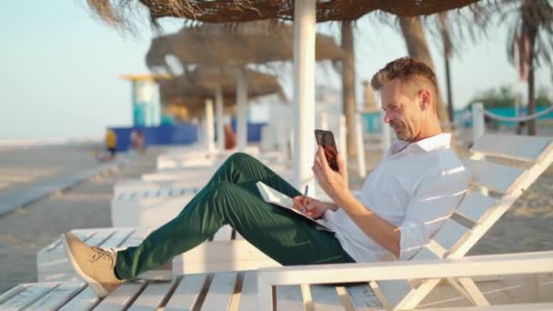 夏日坐在海滩躺椅上 穿着时髦休闲装的成熟男性经理一边一边一边用手机讲话 一边在规划师的记事本上记笔记 — 图库视频影像