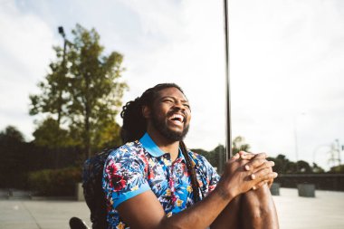 Mutlu sakallı Afrikalı Amerikalı erkek. Rastalı saçlar ve günlük kıyafetlerle modern binanın yakınında otururken gözlerini kaçırıyor.