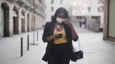 Koruyucu maskeli genç Afrikalı Amerikalı bir kadının akıllı telefonuyla sokakta yürüdüğü görüntüler. 