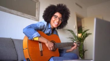 Gülümseyen, kısa kıvırcık saçlı, gözlüklü, rahat koltukta oturup şarkı söyleyen ve dizüstü bilgisayarın önünde gitar çalan Afrikalı Amerikalı bayan müzisyen.