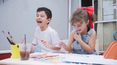 Masada renkli suluboya boyalı boya fırçaları ve beyaz tahtalı malzemelerle dolu kağıtlar taşıyan pozitif çocuklar.