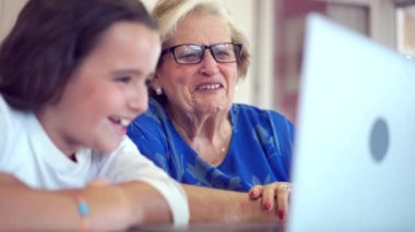 Gülümseyen torun ve büyükanne masada oturuyor ve evdeki aydınlık mutfakta dizüstü bilgisayarda videolar izliyorlar.