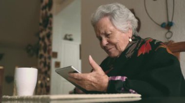 Sıcak giysiler içinde gülümseyen yaşlı bir kadın tablet ve çay bardağıyla masaya oturmuş ekrana bakıyor.