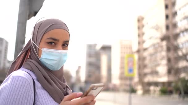 晴れた日に街の路上に立っている民族女性のスカーフや保護マスクを身に着けていると彼女のモバイルの画面を見ての側面図 — ストック動画