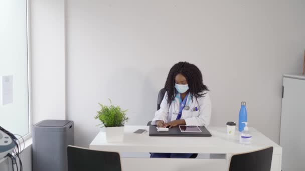 在现代科罗纳韦大流行期间 身穿医用制服 戴防护面罩 头戴平板电脑 在笔记本上书写处方的黑人阳性患者在现代诊所工作时 — 图库视频影像