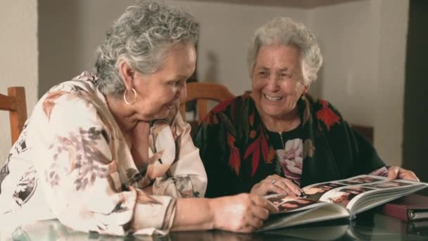 Старшие Сестры Осматривают Фотографии Фотоальбоме Обсуждают Воспоминания Сидя Столом Дома — стоковое видео