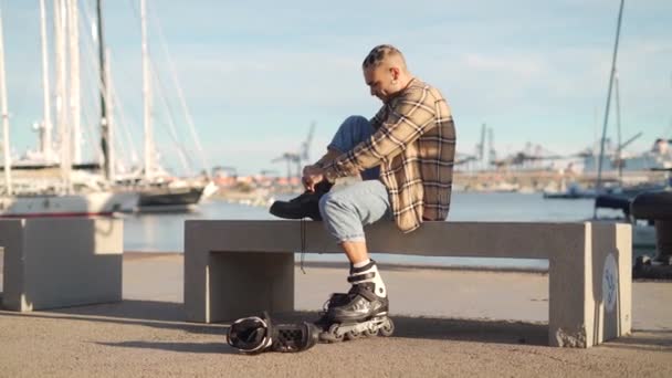 ベンチに座り 余暇時に乗るためのウォーターフロントにローラースケートを置く奇妙な男性の全身 — ストック動画