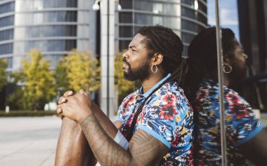 Ciddi, düşünceli, sakallı, rastalı ve sıradan giysileri olan, modern binanın yakınında otururken kameraya bakan şehirli Afrikalı Amerikalı bir erkeğin yan görüntüsü.