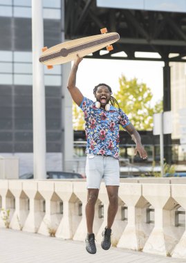 Heyecanlı Afro-Amerikan erkek, kablosuz kulaklıklı, elinde uzun panoyla şehrin kaldırımlarında zıplıyor.