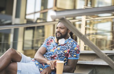 Kablosuz kulaklıklı, boynunda kablosuz kulaklıklar olan düşünceli Afro-Amerikan erkek. Binanın yanındaki merdivenlerde sıcak içecekle otururken mesafeye bakıyor.