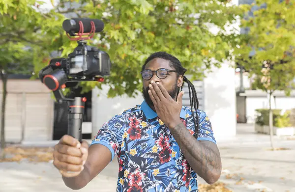 アフリカ系アメリカ人ブロガー 眼鏡でドレッドロック 街の木で街のプロのカメラでビデオを撮影しながらエアキスを送信 — ストック写真