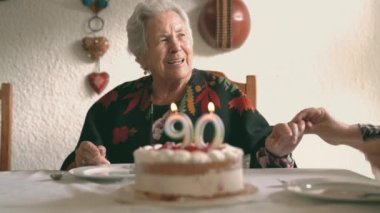 Gri saçlı yaşlı bir kadın ve yaşlı bir kadın yemek masasında oturmuş 90 'ıncı yaş gününü mumlu lezzetli bir pastayla kutluyorlar.
