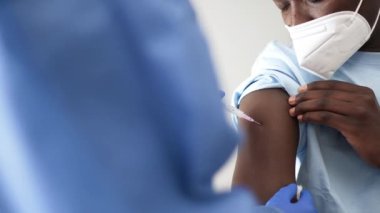 Koruyucu üniformalı ve lateks eldivenli kadın doktor Coronavirus salgını sırasında klinikte Afrikalı Amerikalı erkek hastayı aşılıyor.