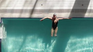 Kapalı havuzun kenarındaki kaldırımda siyah bir mayo giymiş, suda yüzen ve dalmaya giden güzel bir kadın.