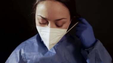 Tıp üniformalı genç bir kadın klinikte siyah arka planda dururken yüz maskelerini çıkarıyor.