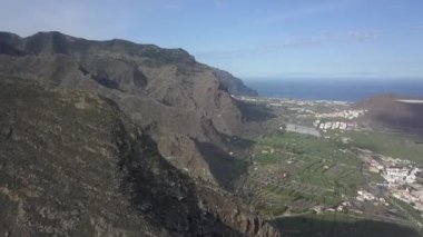 Güneşli bir günde Tenerife 'de kayalık dağ sırasının ve deniz kenarındaki kıyı şehrinin muhteşem drone görüntüsü