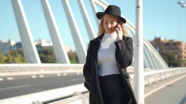 Sırt çantalı ve şapkalı genç sarışın kadın güneşli bir günde asfalt yolda yürürken cep telefonuyla konuşuyor.