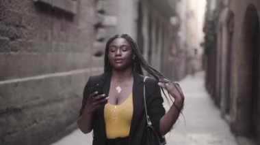 Siyah takım elbiseli genç Afro-Amerikan bir kadının akıllı telefonuyla sokakta yürüdüğü görüntüler. 