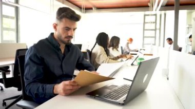Yoğunlaşmış erkek çalışan, bulanık iş arkadaşlarıyla geniş bir ofiste çalışırken dizüstü bilgisayarla masada oturuyor ve belgeleri okuyor.