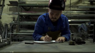 Sert şapkalı, ciddi sakallı bir erkek işçi. Not defterine, hafif garajdaki metal yapıların yanındaki masada kalemle not alıyor.