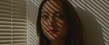 Gölgelerde poz veren sıradan giyinmiş genç ve güzel bir kadın ve kameraya bakan pencere perdeleri.