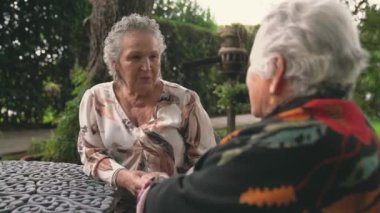 Yaşlı kadın gülümseyip el ele tutuşurken egzotik bir bahçede palmiyeler ve çalılarla oturmuş yaşlı bayan arkadaşınla sohbet ederken.