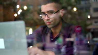 Menekşe gömlekli ve gözlüklü ciddi bir İspanyol erkek yönetici pencerenin arkasındaki masada otururken ve kafede proje üzerinde çalışırken netbook klavyesinde daktilo çalıyor.