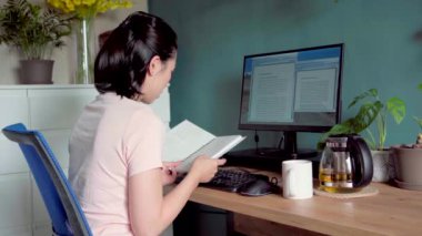 Bilgisayarla masada otururken ve evde proje üzerinde çalışırken not defterinden notlar okuyan Asyalı kadın serbest yazar yan görünümü