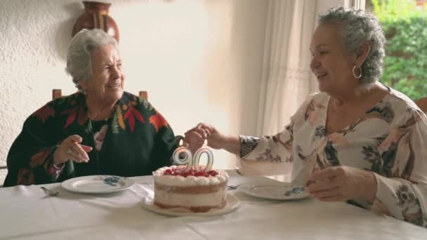 灰色の髪と高齢女性がダイニングテーブルに座っているとろうそくとおいしいケーキで90歳の誕生日を祝います — ストック動画