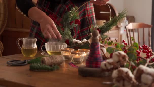 家の居心地の良い部屋で紅茶とろうそくとテーブルに立って休日の祝いのための装飾的なクリスマスリースを作る顔のない創造的な女性のクロップ映像 — ストック動画