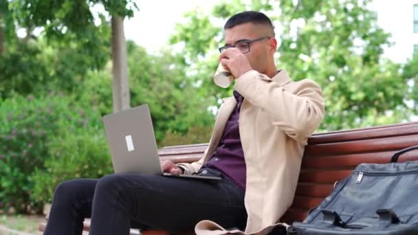 都会の公園のベンチに座っている間にノートパソコンで働くテイクアウトコーヒーのカップとスタイリッシュな衣装やメガネで若いビジネスマンに焦点を当て — ストック動画