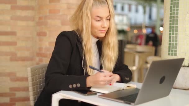 ラップトップとコーヒーでテーブルに座ってカフェで働いている間 プランナーでメモを取る集中した女性フリーランサー — ストック動画