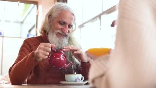 满意的老年男子 留着灰胡子 在自助餐厅里与无名女子交谈时 把刚泡好的茶倒进杯子里 — 图库视频影像