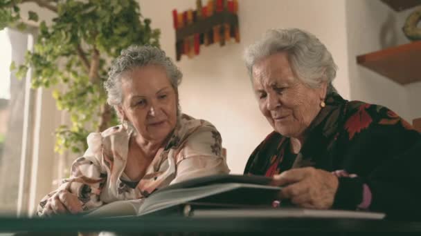 Старшие Сестры Осматривают Фотографии Фотоальбоме Обсуждают Воспоминания Сидя Столом Дома — стоковое видео