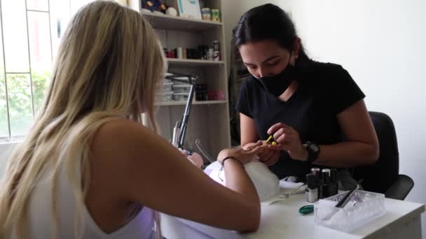 ビューティーサロンで匿名のクライアントの爪に黄色のポーランド語を適用する無菌マスクのエスニック女性マスター — ストック動画
