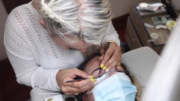 从上面的美容师与镊子应用假睫毛延长病人的眼睛在客厅的保护面具在围观19大流行病 — 图库视频影像