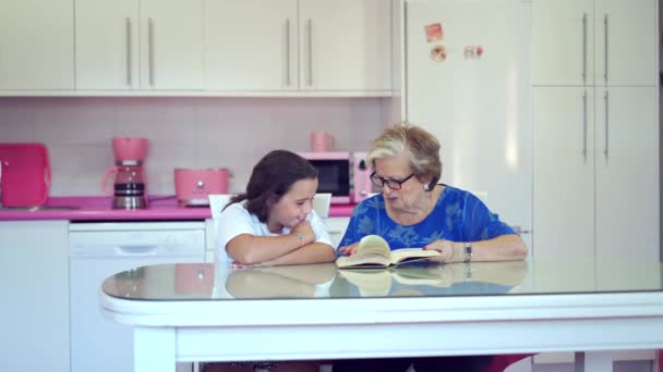 集中祖母でカジュアル服や眼鏡で座ってテーブルと読書本とともに陽気な孫娘とともに台所に自宅 — ストック動画
