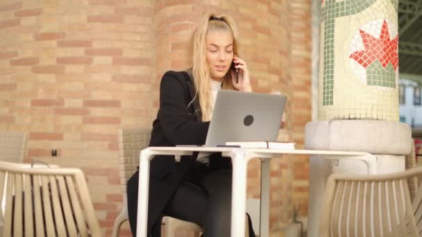 カフェのベランダのテーブルでリモートで座っている間 携帯電話で話している若い女性フリーランサー — ストック動画