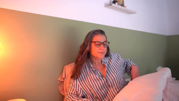 ベッドサイドランプを回すストライプのスリープウェアを身に着けている中年女性のリアルタイムは 朝の寝室で寝ている間に興味深い本を読み始める — ストック動画