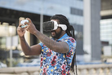 African American male with dreadlocks in contemporary VR goggles surfing cellphone while exploring virtual reality on street with modern buildings clipart