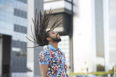 Kentteki modern binalarla caddede dikilirken siyah rasta giymiş, saçlarını sallayan Afro-Amerikan erkeklerin yan görüntüsü.