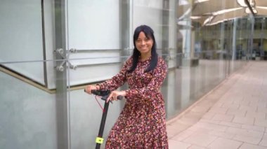 Gülümseyen, uzun siyah saçlı, elektrikli mobiletiyle ofis binasında duran ve kameraya bakan etnik kadına yakınlaş.