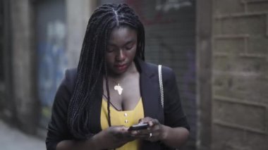 Siyah takım elbiseli genç Afro-Amerikan bir kadının akıllı telefonuyla sokakta yürüdüğü görüntüler. 