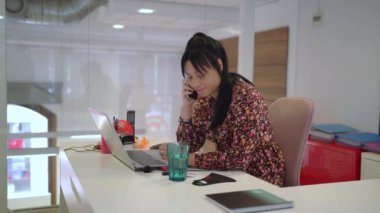 Etnik kadın yöneticinin cep telefonuyla konuşma ve modern çalışma alanında dizüstü bilgisayarda yazma görüntüsünü yakınlaştır