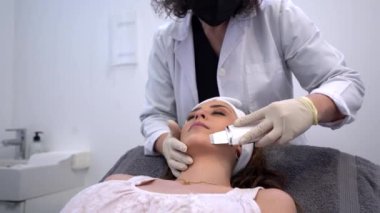 Tanımlanamayan kozmetik uzmanı, ultrasonik yüz derisi yüzüyor. Modern güzellik kliniğinde tıp masasında uzanan rahat bir kadın için.