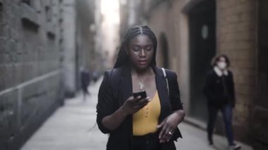 Siyah takım elbiseli genç bir Afro-Amerikan kadının akıllı telefonuyla sokakta yürüdüğünü gösteren görüntüler. 